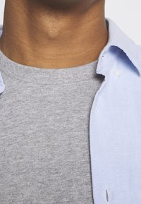 Světle modrá košile na knoflíky částečně překrytá šedým svetr s kulatým výstřihem, s viditelnými texturami látky a detaily límce.