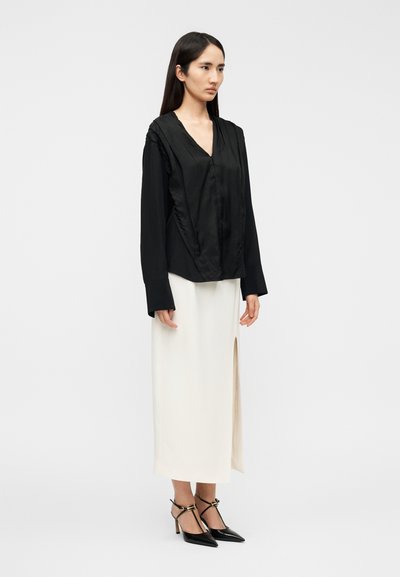Μπλούζα από μαύρο σατέν με βαθύ V-λαιμό και βολάν στους ώμους, σε συνδυασμό με κρεμ φούστα με πλευρικό σκίσιμο και μαύρες μυτερές γόβες.