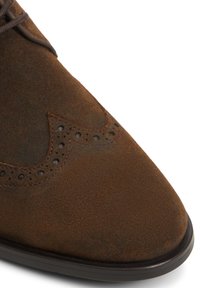 Zapato de ante marrón con punta afilada, detalles decorativos de perforaciones en la puntera y cordones marrones. Presenta un acabado texturizado y suela negra.