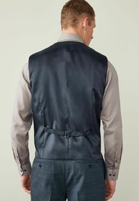 Navy satin vestillä on sileä rakenne, jossa on takana solki ja säätöremmi, yhdistetty kevyen harmaan paidan ja teksturoidun sinisen housun kanssa.