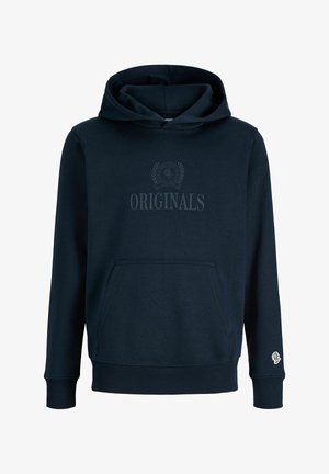 Garçon aux cheveux bouclés porte une veste beige sur un sweat à capuche noir, un jean bleu large et des chaussures beiges, debout devant un fond blanc uni.