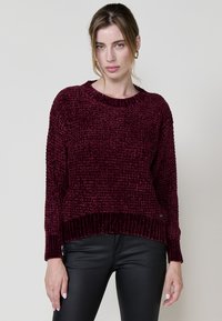 Burgunderfarbener Strickpullover mit strukturiertem Muster und gerippten Bündchen. Rundhalskragen und lässige Passform. Kombiniert mit schwarzen engen Hosen.