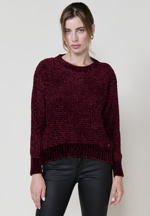 Pull en maille bordeaux avec motif texturé et poignets côtelés. Col rond et coupe décontractée. Associé à un pantalon noir ajusté.