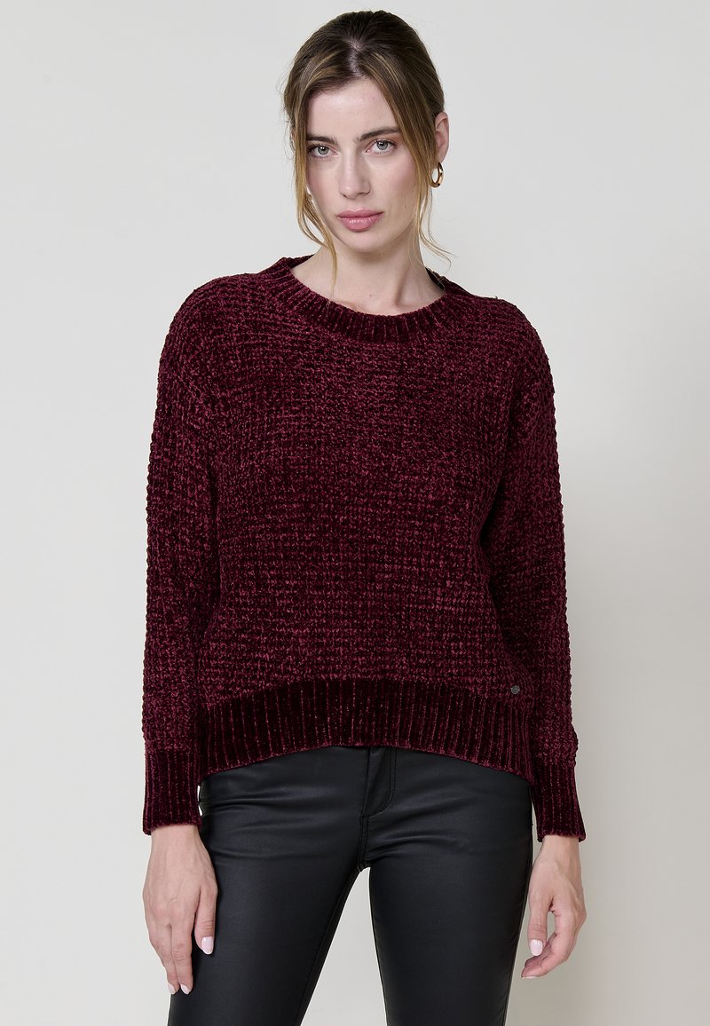 Burgunderfarbener Strickpullover mit strukturiertem Muster und gerippten Bündchen. Rundhalskragen und lässige Passform. Kombiniert mit schwarzen engen Hosen.