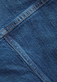 Tume sinine denimkangas tekstuuriga pinnaga, millel on kontrastsed heledat pruunid õmblused õmbluste ja servade ääres, rõhutades vastupidavust.