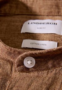 Colletto di camicia in cotone-lino marrone con un bottone bianco e etichette con la scritta "Lindbergh" e "Slim Fit", che mostrano un tessuto strutturato.