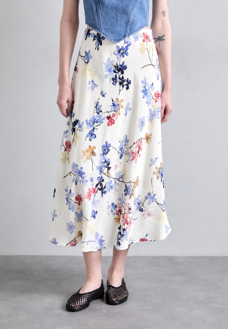 Floral midi skirt in cream, adorned with blue, red, and yellow blossoms. Lightweight fabric with a flowing, relaxed fit. Black mesh slip-on shoes.