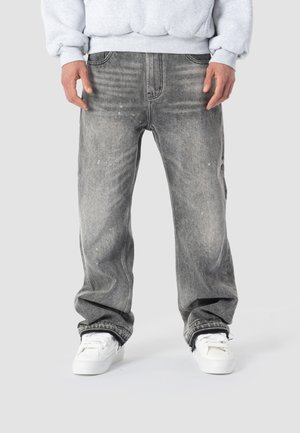 Person trägt verwaschene schwarze, locker sitzende Jeans mit ausgefransten Säumen und weißen Turnschuhen, kombiniert mit einem hellgrauen Sweatshirt.