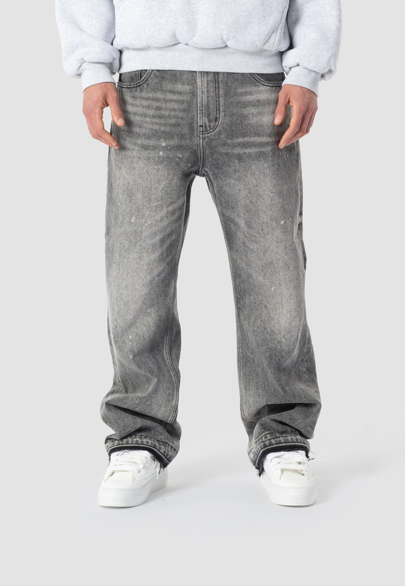 Persona che indossa jeans neri sbiaditi a vestibilità larga con orli sfrangiati e sneakers bianche, abbinati a una felpa grigio chiaro.