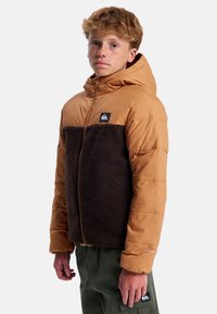 Brązowa i beżowa pikowana kurtka z kapturem z polaru z przodu, zapinana na zamek błyskawiczny, z naszywką z logo na lewej piersi. Teksturowana powierzchnia na polarze.