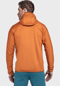 Giacca arancione con cappuccio e texture a coste, caratterizzata da un collo alto e maniche raglan. Indossata sopra pantaloni corti blu, che mostrano uno stile casual.