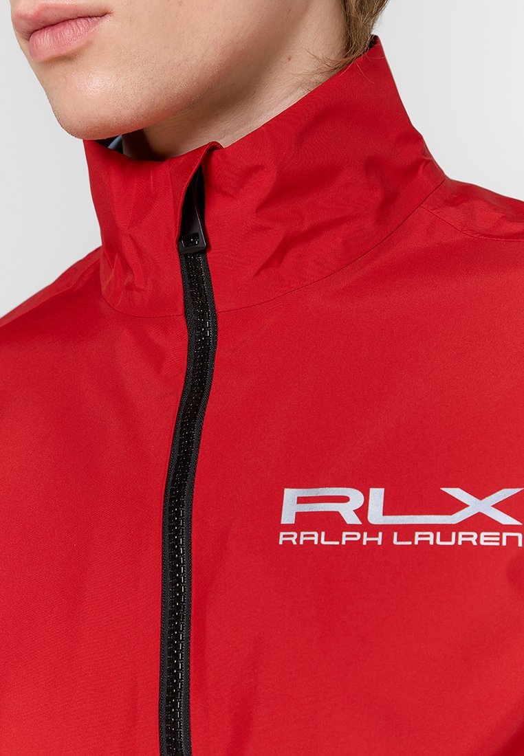 Chaqueta de lluvia roja con alto cuello, con cremallera negra y texto blanco "RLX RALPH LAUREN." Hecha de material suave y ligero.