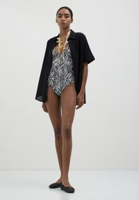 Camicia nera oversize sopra un costume da bagno a motivi bianco e nero, completata da una collana di colore chiaro e da ballerine nere a fantasia.