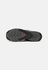 Semelle de chaussure de randonnée noire avec un motif de traction robuste, présentant de profonds sillons et un accent rouge, conçue pour une adhérence sur divers terrains.
