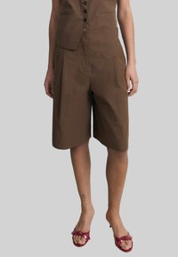 Brown, knee-length shorts with a tailored fit, featuring pleats and side pockets. Made from smooth fabric. Paired with red heeled sandals.