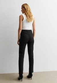 Pantalones de mezclilla negros con cintura alta, corte ajustado y bolsillos traseros, combinados con una blusa sin mangas blanca y botines de tacón negros.