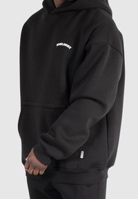 Sweat à capuche noir en tissu doux. Comprend une poche avant, des poignets côtelés et un logo blanc sur la poitrine. Design simple et coupe décontractée.