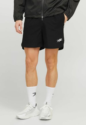 Homme portant un short de sport noir avec un logo blanc, des chaussettes blanches avec logo, des chaussures de course blanches et une veste noire, se tenant devant un fond uni.