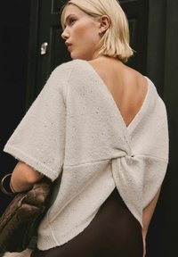 Blonde woman wearing a white short-sleeve knit top with a deep V-back and twist detail, holding a brown woven handbag.