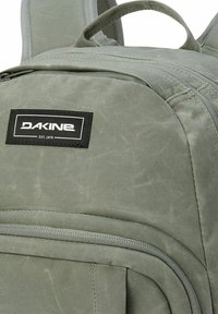 Zaino in tessuto verde con superficie texturizzata, dotato di una patch logo nera e una tasca con zip. Maniglia superiore robusta e spallacci regolabili.