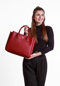 Rote Ledertasche mit flachen Griffen, quadratischer Form, glatter Textur und einem Logo-Accento auf der Vorderseite. Getragen von einer Person in schwarzer Kleidung.