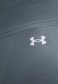 Κοντινή εικόνα από γκρι ύφασμα Under Armour με ορατές ραφές και λευκό λογότυπο Under Armour σε λείο αθλητικό υλικό.