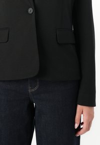 must lääne kuub mustade flap taskutega, kantud tumesiniste teksade kohal, üks käsi rahulikult külje peal.