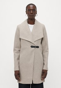 Beige wollen jas met een brede kraag, asymmetrische sluiting en een zwart gesp-detail. Recht afgesneden mouwen en een jurkachtige lengtesilhouet.