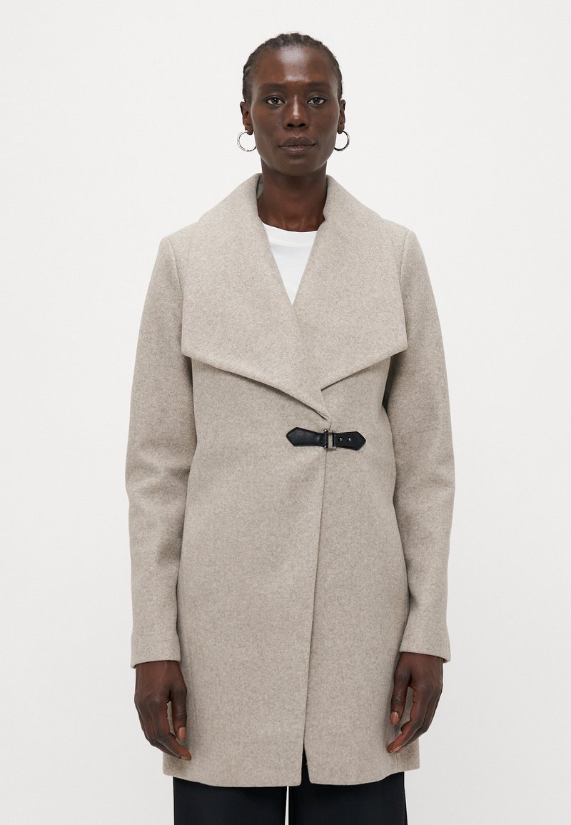 Manteau en laine beige avec un large col, fermeture asymétrique et détail en boucle noire. Manches droites et silhouette mi-longue.
