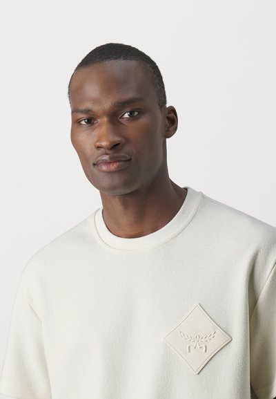 Kreemivärvi sweatshirt, millel on tekstuuriga kangas, ümmargune kaelus ja tõstetud rombikujuline logopatch vasakul rinnal.
