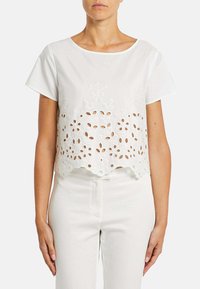 Hvid cropped bluse med korte ærmer, prydet med blomsterbroderi og udskæringer langs kanten. Glat tekstur, minimalistisk design.