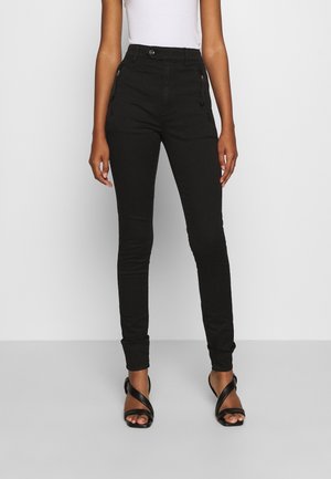 Pantalon noir ajusté taille haute avec fermeture à bouton et fermeture éclair, porté avec des talons noirs à brides et un haut blanc sans manches.