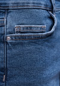 Κοντινό πλάνο τσέπης μπλε τζιν με ορατές ραφές και μεταλλική τρουκ σε υφή υφάσματος.