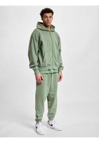 Haut à capuche zippé vert clair avec poches avant, associé à un pantalon de sport assorti. Baskets blanches avec des accents noirs. Le tissu semble doux et décontracté.