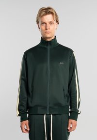 Hombre rubio con chaqueta deportiva verde oscuro con rayas crema en las mangas y pantalones a juego, de pie contra un fondo gris liso.