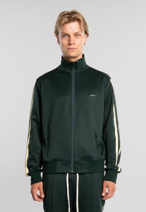 Blondýnový muž v tmavě zelené sportovní bundě s krémovými pruhy na rukávech a odpovídajících kalhotách, stojící před jednobarevným šedým pozadím.