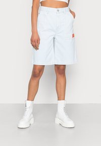 Shorts de mezclilla de color azul claro con bolsillos delanteros y un parche de logo rojo, combinados con botas de tobillo blancas con cordones sobre un fondo neutro.