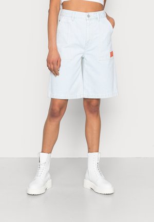 Shorts de mezclilla de color azul claro con bolsillos delanteros y un parche de logo rojo, combinados con botas de tobillo blancas con cordones sobre un fondo neutro.
