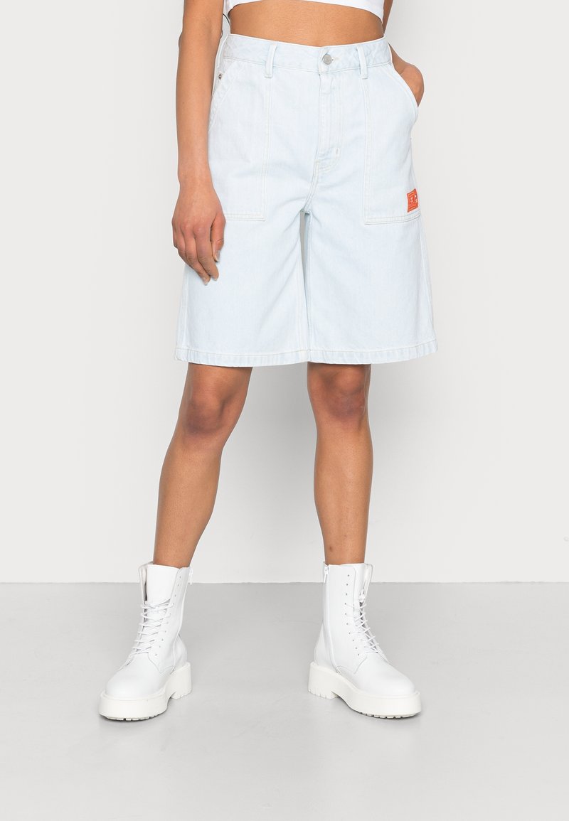 Shorts de mezclilla de color azul claro con bolsillos delanteros y un parche de logo rojo, combinados con botas de tobillo blancas con cordones sobre un fondo neutro.