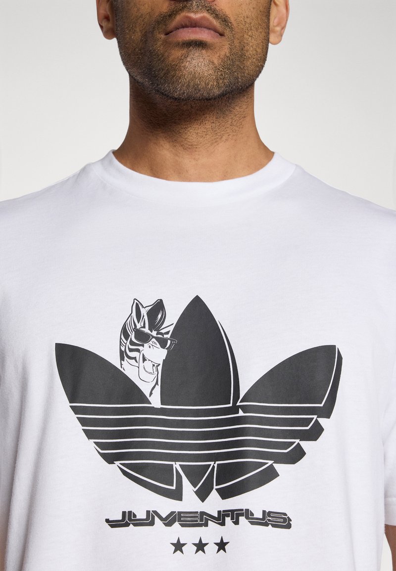 Weißes T-Shirt mit einer schwarzen Illustration eines Zebras mit Sonnenbrille, das über einem stilisierten Blättermotiv mit gestreiften Details und Text liegt.