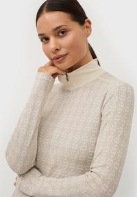 Beige langesleeve top met een hoge kraag, voorzien van een gestructureerd geometrisch patroon en een ritssluiting aan de voorkant bij de nek.