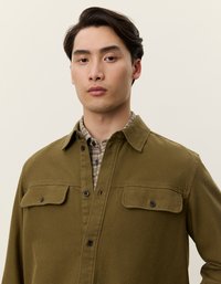 Κυπαρισσί καμπαρντίνα από κοτλέ με κουμπωτή μπροστινή πλευρά, δύο τσέπες στο στήθος και κλασικό γιακά, φορεμένη πάνω από πουκάμισο με καρό σχέδιο.