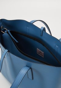 Sininen nahkainen olkalaukku, jossa on kaksinkertaiset kahvat, varustettu vetoketjullisella sisätaskulla ja merkkilabelillä. Sujuva pinta ja minimalistinen muotoilu.