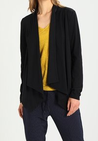 Svart kofta med sjalkrage, öppen framsidan och draperad design, lagd över en gul V-ringad skjorta. Matchas med marinblå randiga byxor.