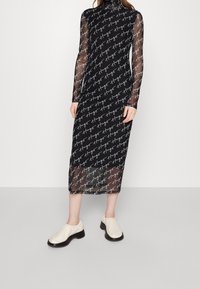 Robe en mesh noire avec un motif textuel blanc répété, col montant, manches longues et silhouette ajustée. Associée à des chaussures crème à enfiler.