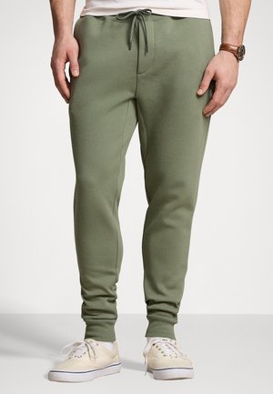 Mann trägt olivgrüne Jogginghose mit Kordelzug in der Taille und weiße Canvas-Sneaker, stehend vor hellgrauem Hintergrund.