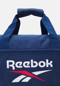 Ναυτικό μπλε τσάντα Reebok με διπλές λαβές, κλείσιμο με φερμουάρ και μεγάλο άσπρο λογότυπο. Διαθέτει κόκκινη διακοσμητική λωρίδα σε γεωμετρικό σχέδιο.