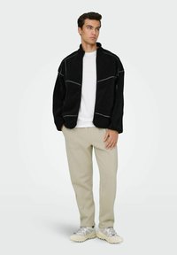 Veste zippée en polaire noire avec des coutures grises contrastantes, portée sur une chemise blanche, associée à un pantalon beige et des baskets de couleur claire.