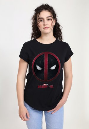 Ung kvinde med mørkt krøllet hår iført en sort Deadpool T-shirt og lyseblå jeans, der står mod en ensfarvet lys baggrund.