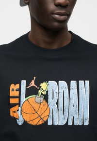 Μαύρο βαμβακερό t-shirt με γραφικό τύπωμα ενός μπάσκετ, πολύχρωμες λεπτομέρειες και το κείμενο «AIR JORDAN» σε γραμματοσειρά με φθορές.